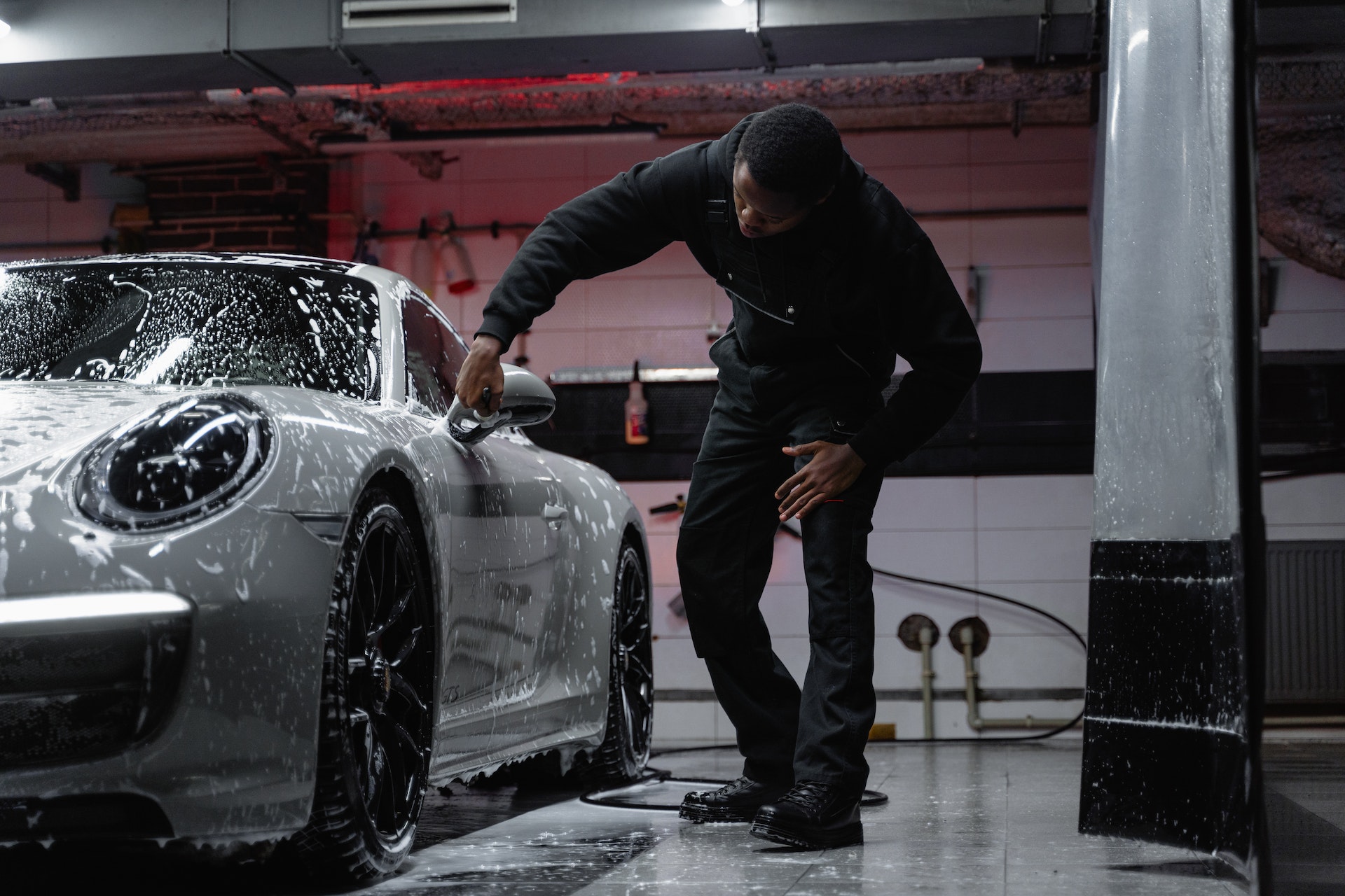 Inside a car detailing garage, a man in black overalls works cleaning a soapy, silver Porsche sports car.