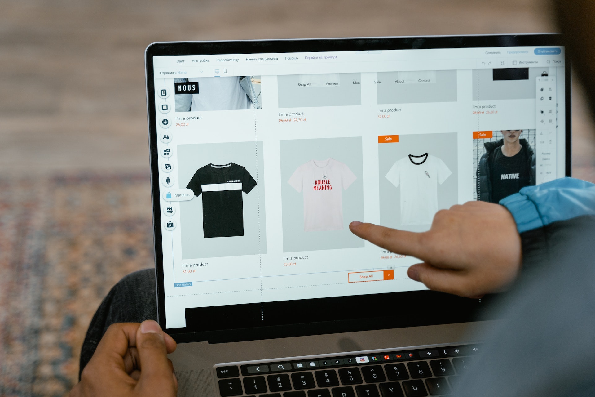 A laptop is open with a browser window displaying a clothing sales website while a hand points at an image of a t-shirt.